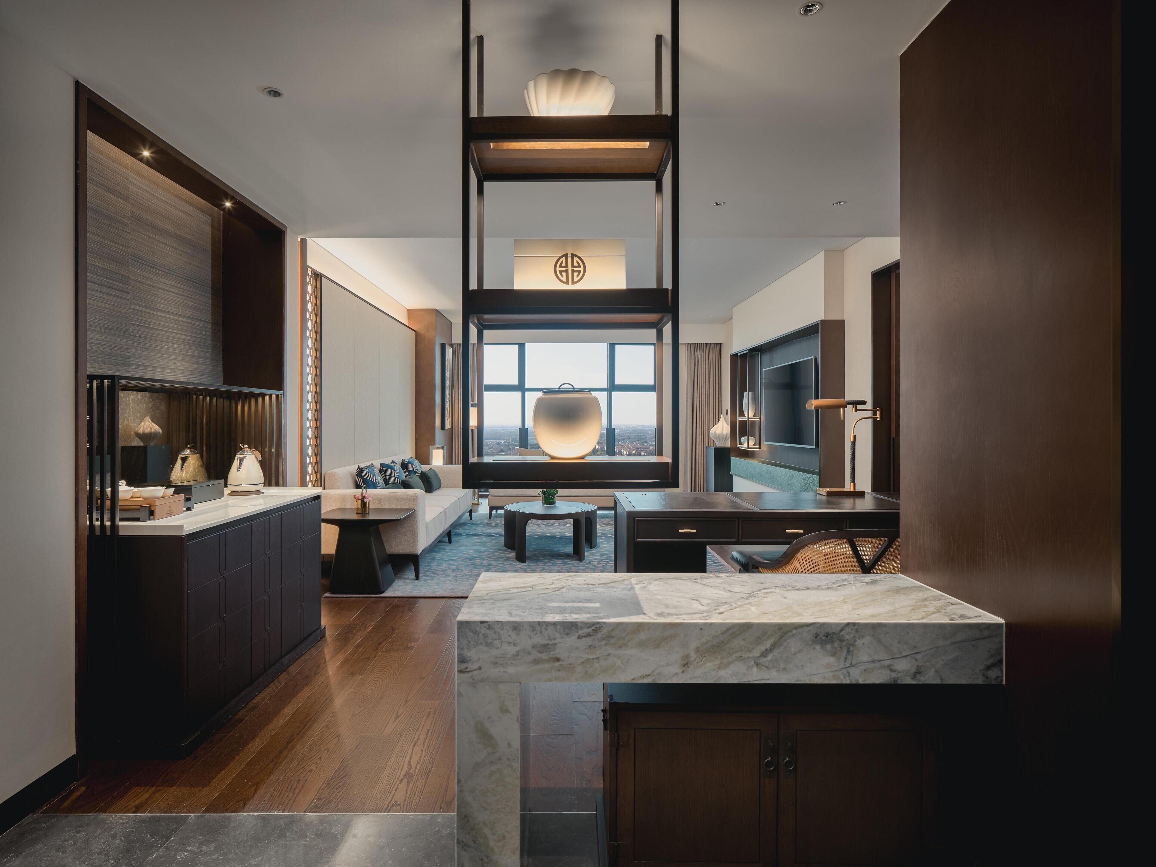 Una habitación de hotel moderna con una isla de cocina de mármol, un sofá, una mesa de comedor y un ventanal con vistas a la ciudad.
