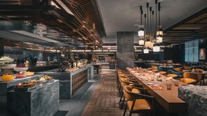 A modern restaurant interior with a long communal dining table, multiple seating areas, and a central bar counter with food displays. - Hualuxe Kunshan Huaqiao (Suzhou)