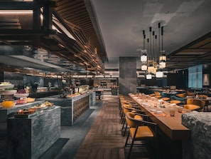 Un restaurant moderne avec un intĂ©rieur Ă©lĂ©gant, une longue table Ă manger commune, plusieurs espaces de dĂ©tente et un bar central oĂč sont exposĂ©s des plats.