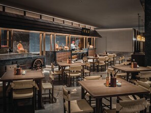 A restaurant with wooden tables and chairs, a counter with a chef, and a large window with blinds. - Hualuxe Kunshan Huaqiao (Suzhou)