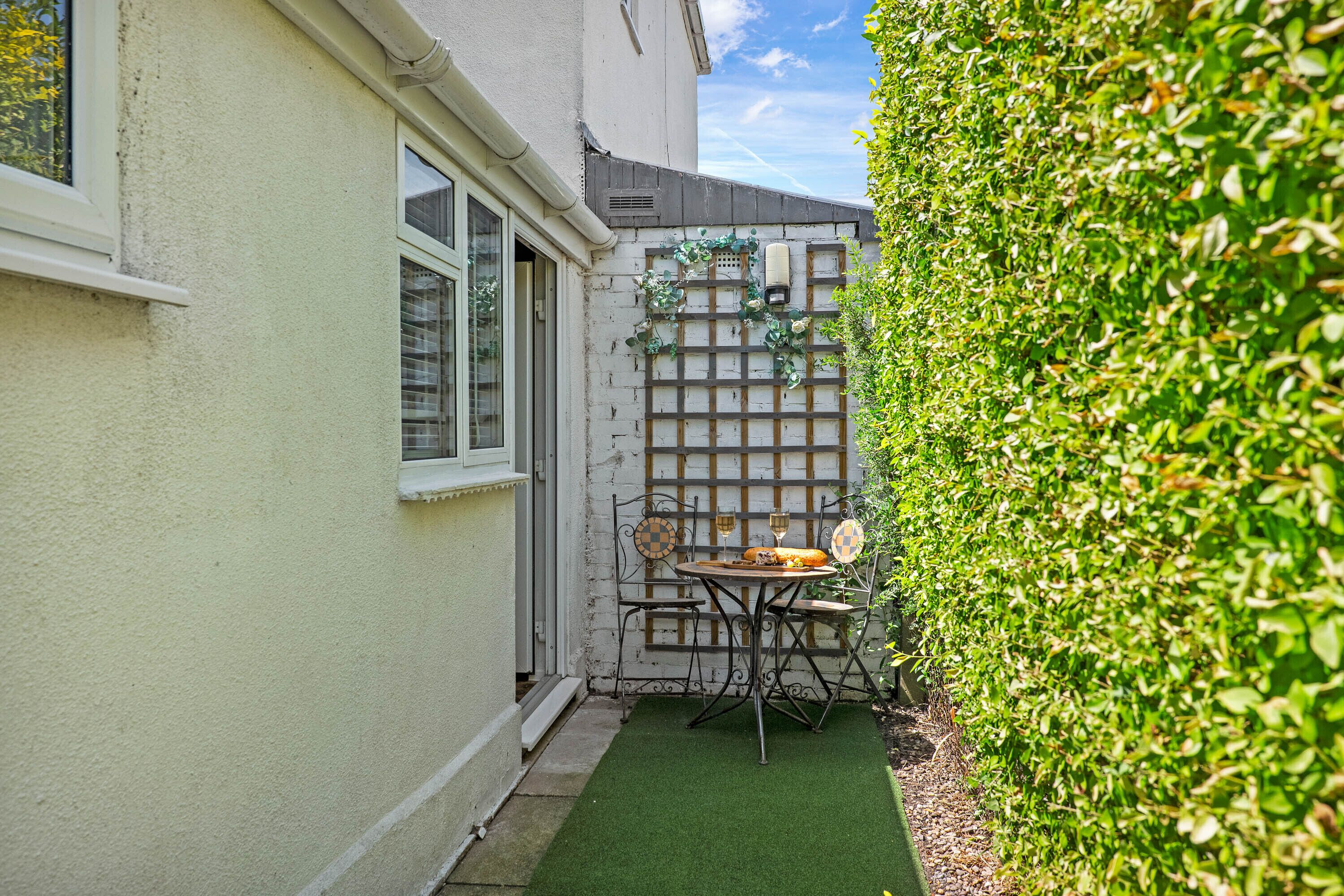 A small patio with a table and chairs, a wooden trellis with climbing plants, and a hedge.