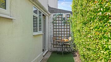 A small patio with a table and chairs, a wooden trellis with climbing plants, and a hedge.