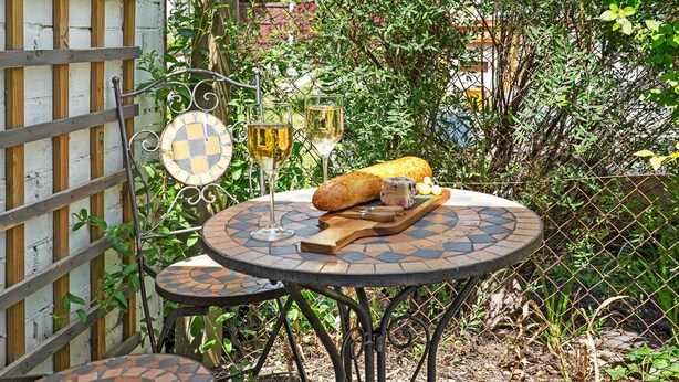 Eine Terrasse mit einem runden Tisch und einem kleineren Tisch, beide mit belegten Fliesen. Auf dem größeren Tisch stehen zwei Weingläser, ein Baguette und ein Schneidebrett mit einem Messer.