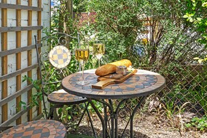 A patio with a round table and a smaller table, both with patterned tile tops. There are two wine glasses on the larger table, a baguette, and a cutting board with a knife.