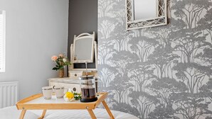 Ein ordentlich eingerichtetes Schlafzimmer mit einem Nachttisch aus Holz, auf dem eine Kaffeemaschine, Tassen und eine Blume stehen. Darüber an der Wand ist ein Spiegel befestigt.