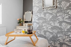 A neatly arranged bedroom with a wooden bedside table holding a coffee maker, cups, and a flower. A mirror is mounted on the wall above the table.