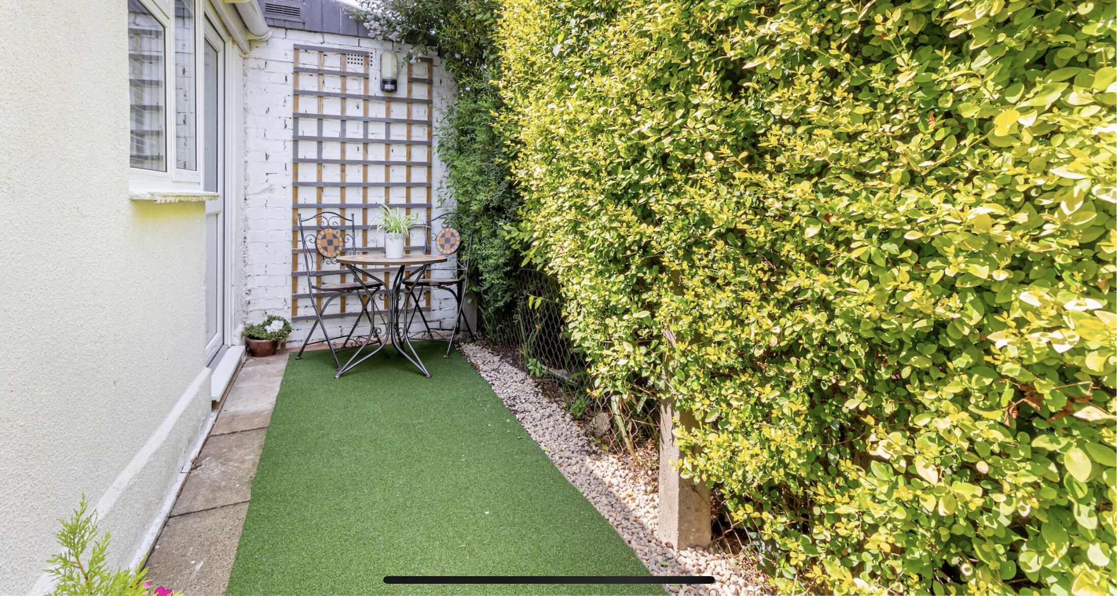 A small outdoor patio with a table and chairs, a potted plant, and a hedge.