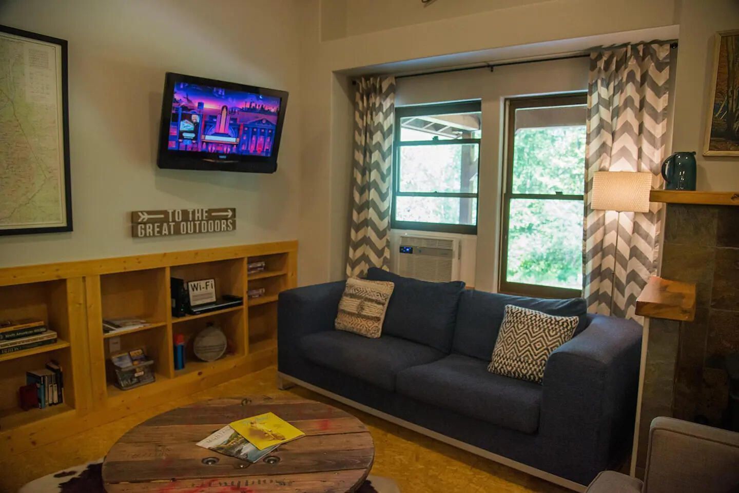 A living room with a blue sofa, a coffee table with magazines, a TV on the wall, and a sign that says "TO THE GREAT OUTDOORS".
