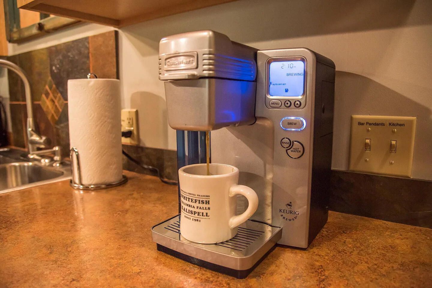 A coffee machine with a digital display, a coffee cup, and a paper towel dispenser on a kitchen counter.