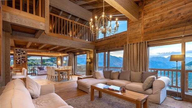 Un vaste salon doté d’un haut plafond, de poutres apparentes, d’un lustre, d’un ensemble de canapés, d’un coin repas et de grandes fenêtres offrant une vue sur les montagnes.