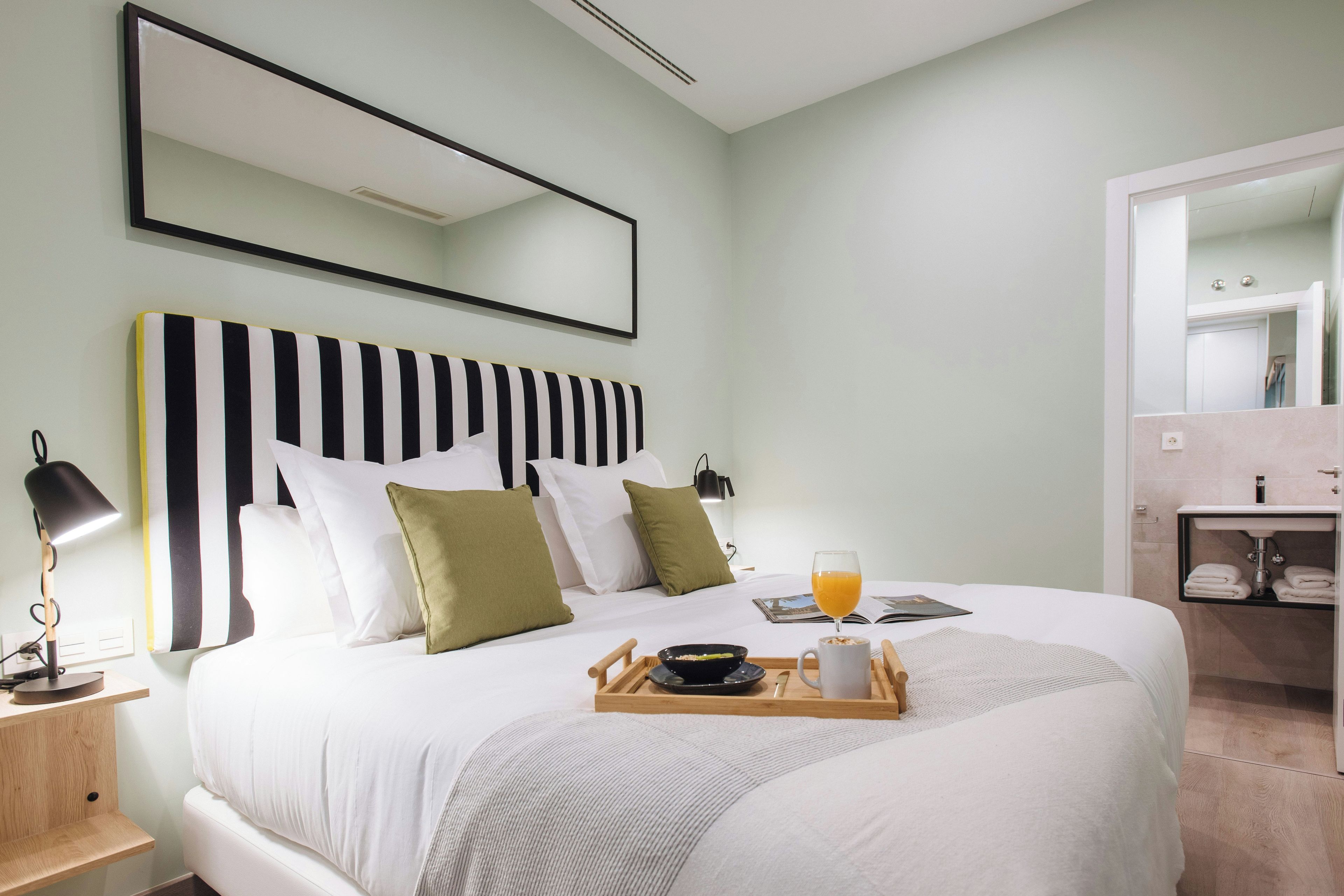 Un lit bien fait, avec un plateau contenant des articles pour le petit-déjeuner, une lampe de chevet et un grand miroir au-dessus du lit.