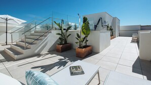 Een terras op het dak met een glazen balustrade, planten in potten, een witte tafel en een trap.