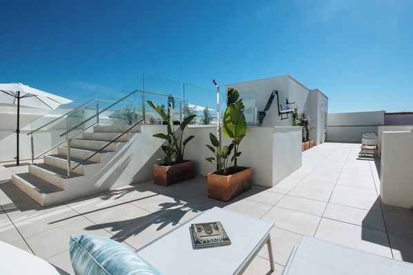 Eine Dachterrasse mit Glastrennwand, Blumentöpfen, einem weißen Tisch und einer Treppe.