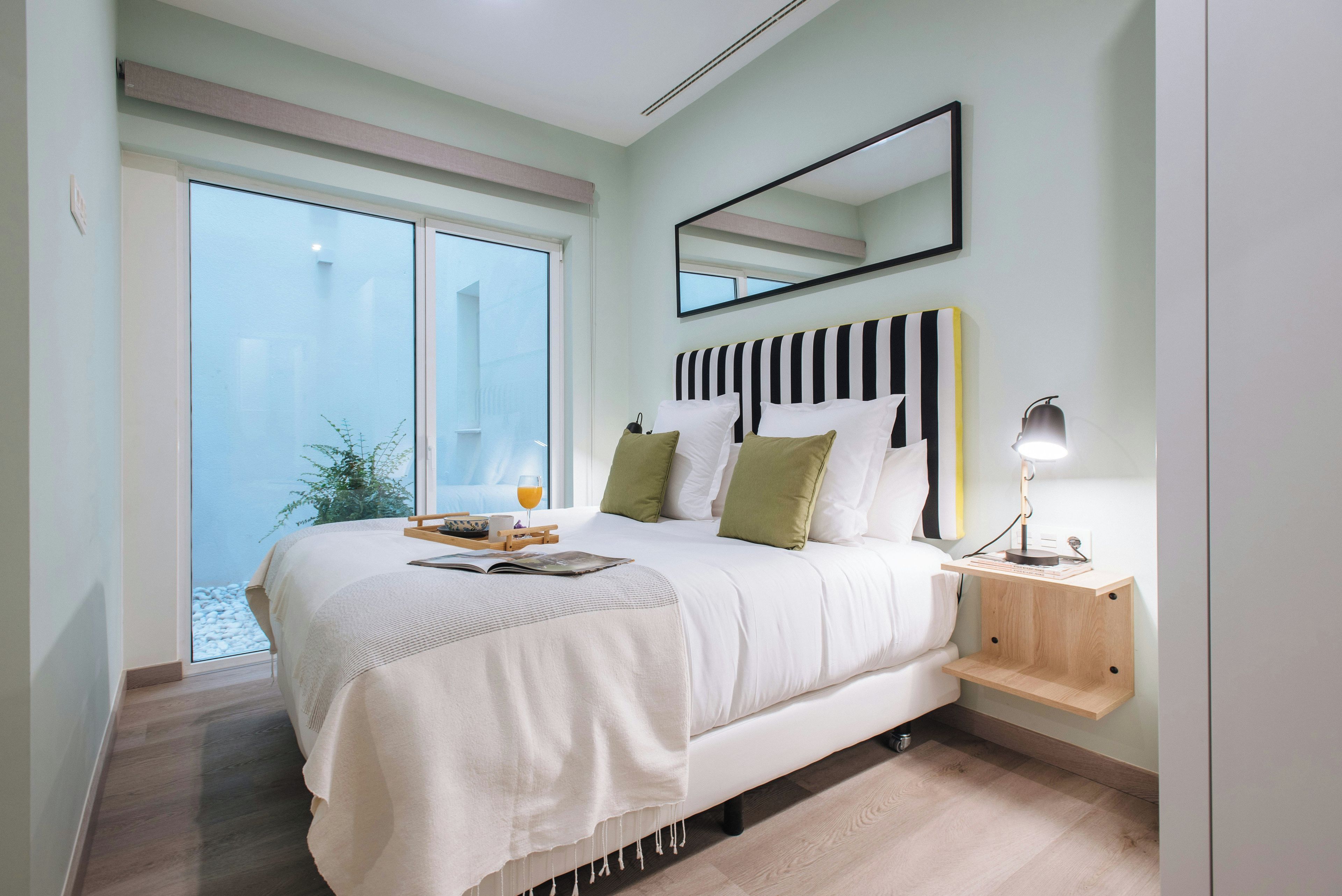 Un dormitorio con una cama grande, una mesita de noche con lámpara, una ventana con vistas y un espejo en la pared.