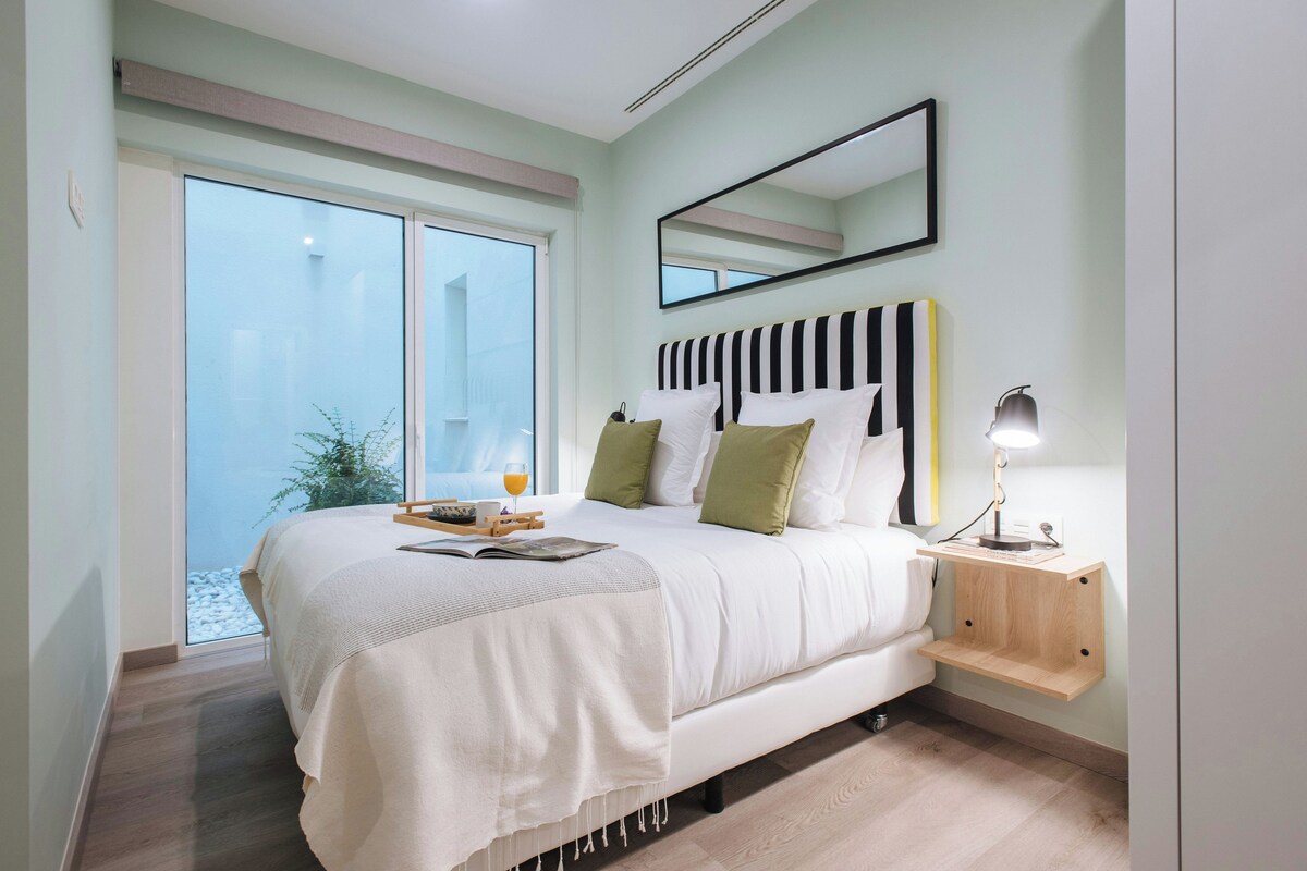 A bedroom with a large bed, a bedside table with a lamp, a window with a view, and a mirror on the wall.