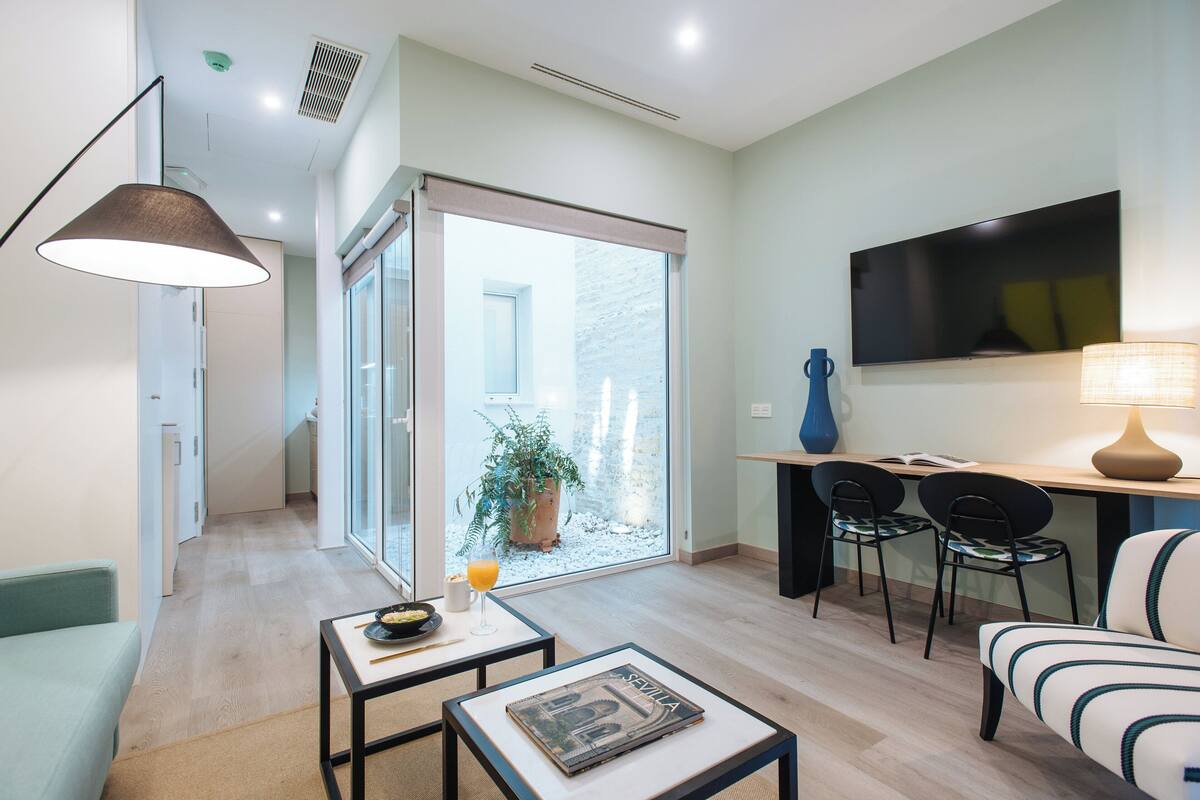 A modern living room with a sofa, a coffee table, a dining area with chairs, a TV, and a sliding glass door leading to a balcony with a potted plant.