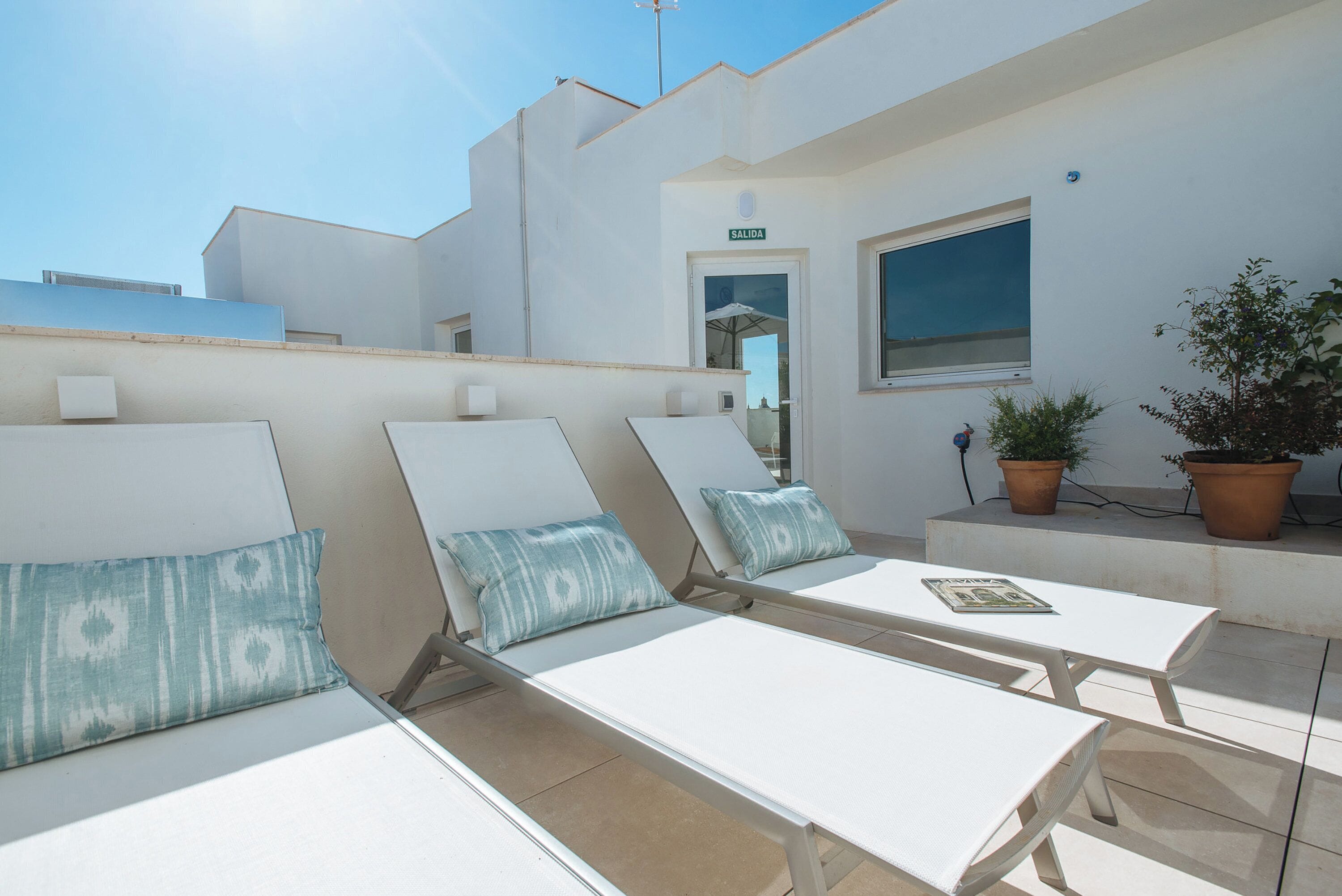 A rooftop terrace with white lounge chairs and a small table, a door labeled "SALIDA," and potted plants.