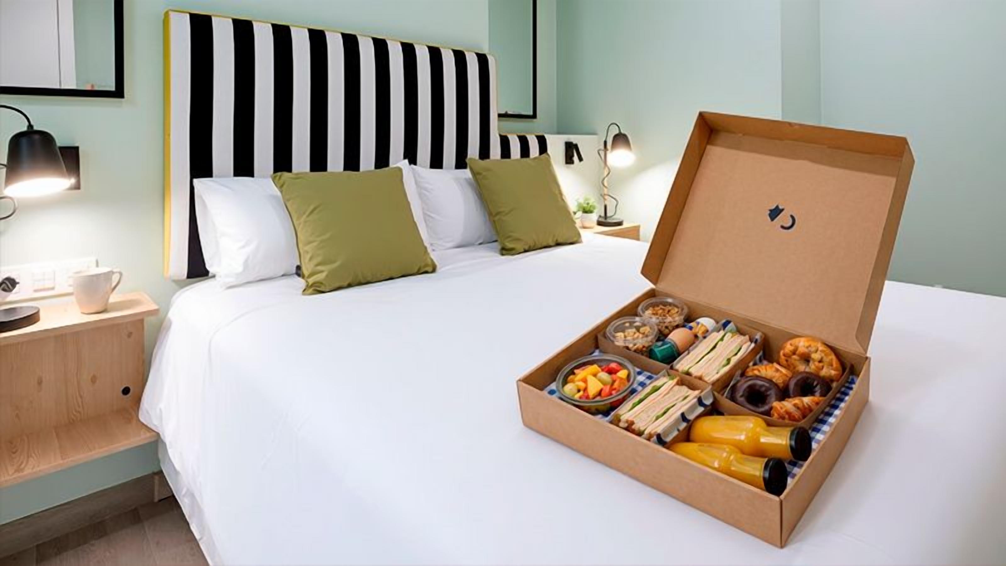 A neatly made bed with a striped headboard, a bedside table with a lamp, and a breakfast tray with various food items.