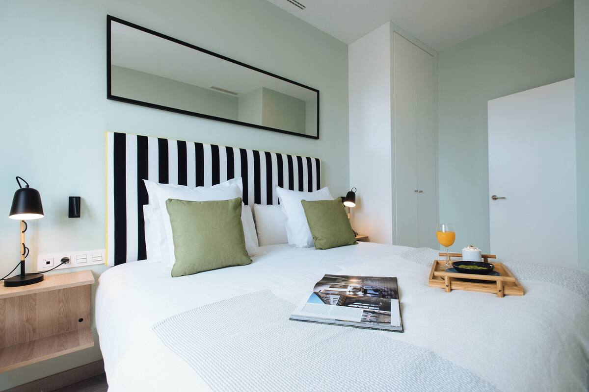 A neatly made bed with a striped headboard, a bedside table with a lamp, and a tray with a glass of orange juice and a book.
