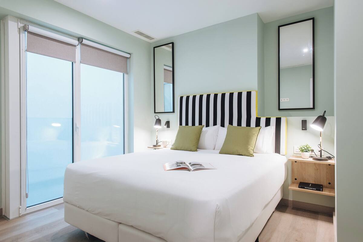 A modern bedroom with a large bed, a nightstand with a lamp, a mirror, and a sliding glass door leading to a balcony.