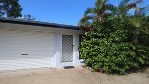 Un bâtiment d’un seul étage, doté d’un portail de garage blanc fermé, d’une petite fenêtre protégée par un moustiquaire et d’un arbuste verdoyant orné de fleurs rouges.
