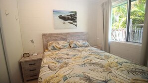 Un dormitorio con cama, mesita de noche, reloj de pared, cuadro y ventana con vista a follaje.