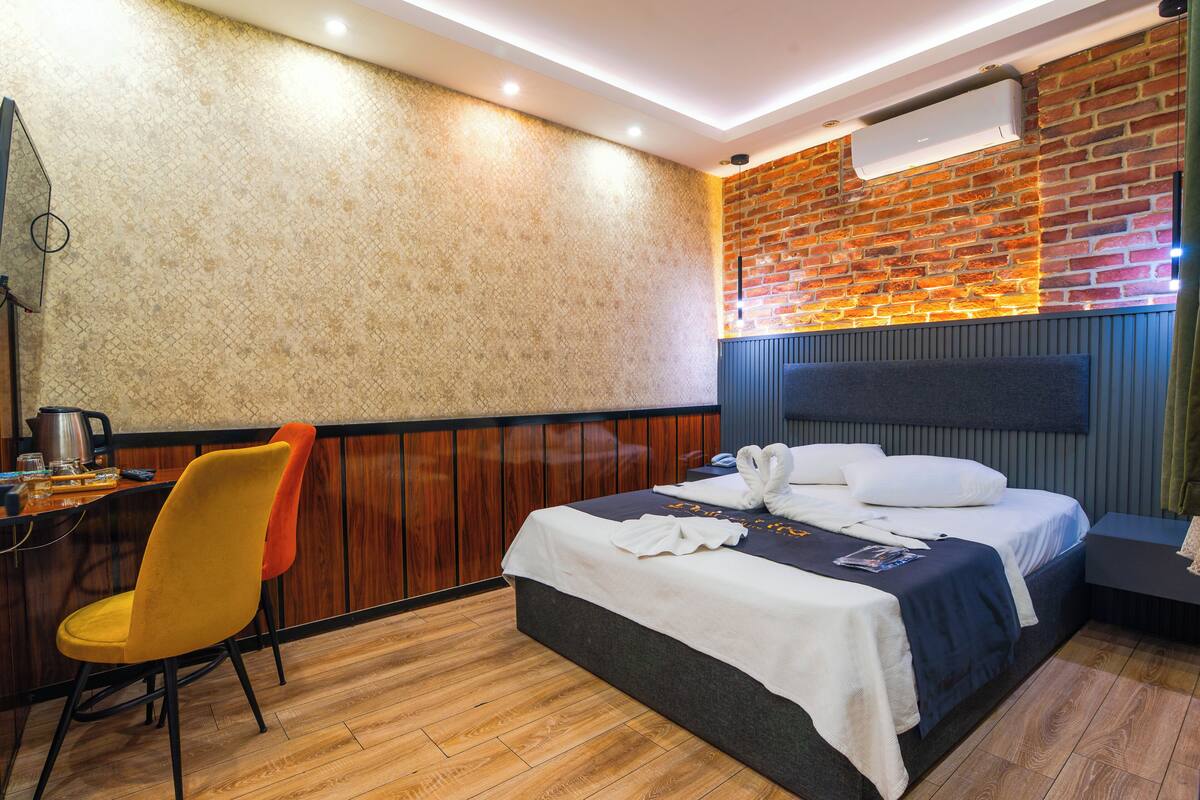 A hotel room with a bed, a wooden chair, a table with a kettle, and a brick wall.