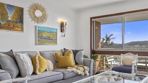 A living room with a grey sofa, yellow cushions, a glass coffee table, and a view of a landscape through a large window.