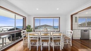 A dining area with a wooden table and white chairs, large windows with blinds, and a view of the ocean.
