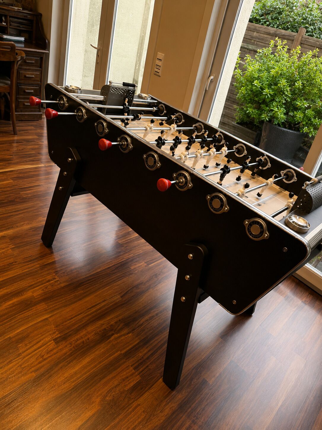 A foosball table in a room with wooden flooring and a view of greenery outside.