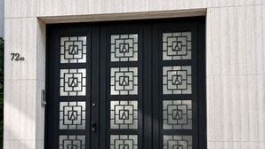 Eine schwarze Doppeltür mit geometrischen Glassmustern und dem Buchstaben 'A' an einer Gebäudefassade.