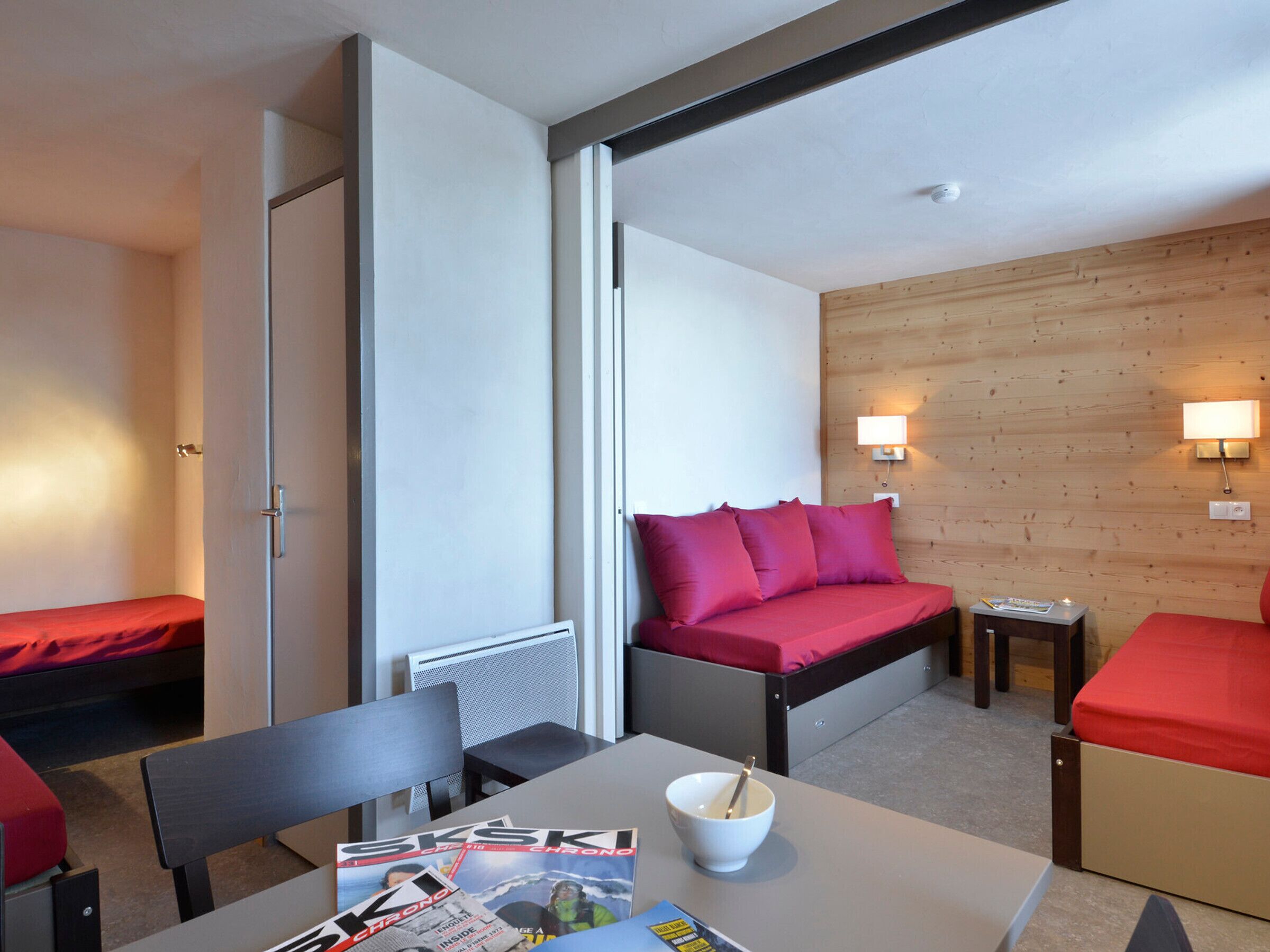A compact room with a bed, a table with a bowl and magazines, and a chair.