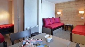 A compact room with a bed, a table with a bowl and magazines, and a chair.