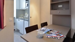 A compact kitchen with white appliances, a dining table with magazines, and a small shelving unit.