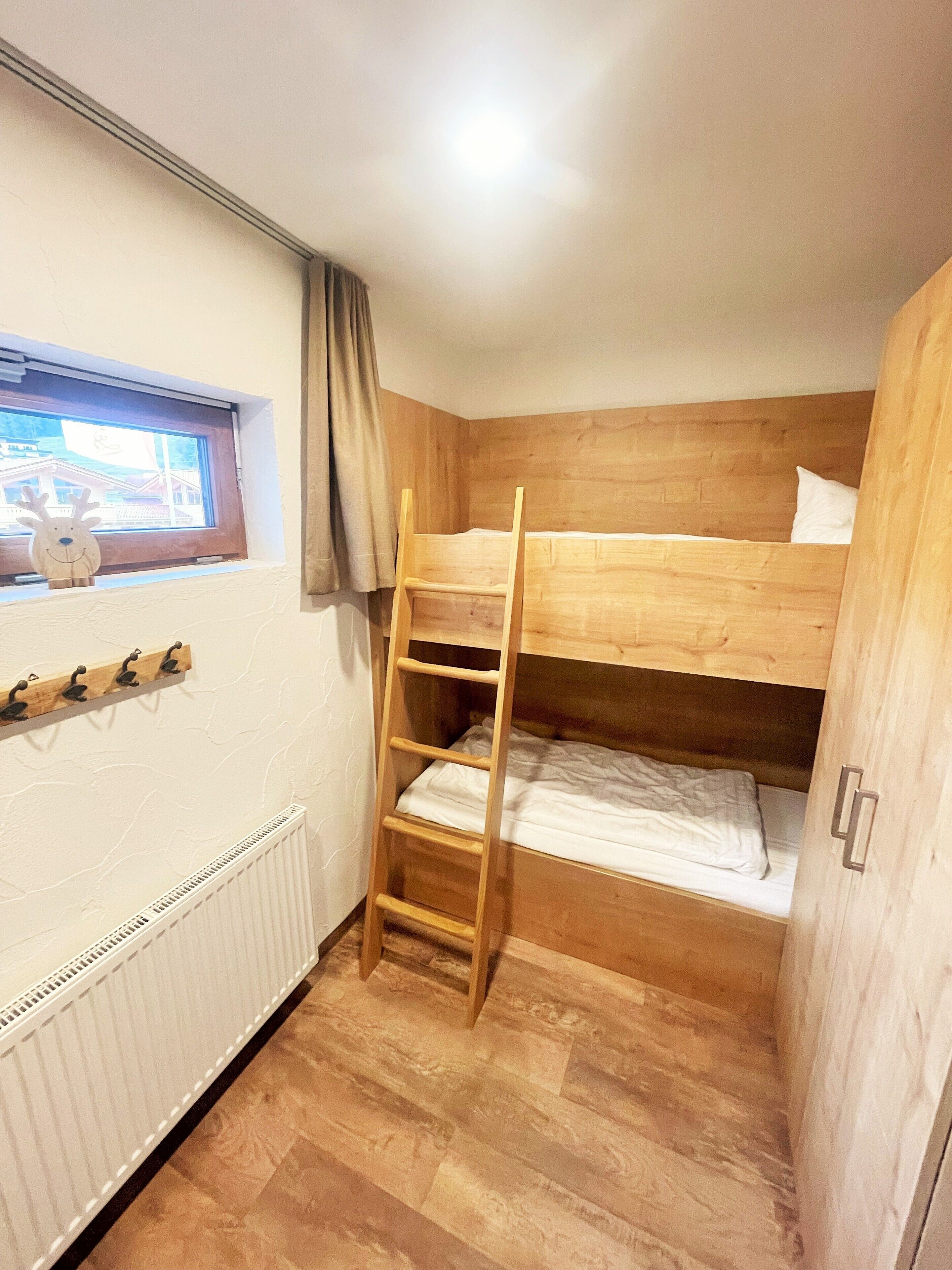 Una habitación pequeña y funcional con una cama litera, una escalera de madera, una ventana que da a un edificio y un radiador.