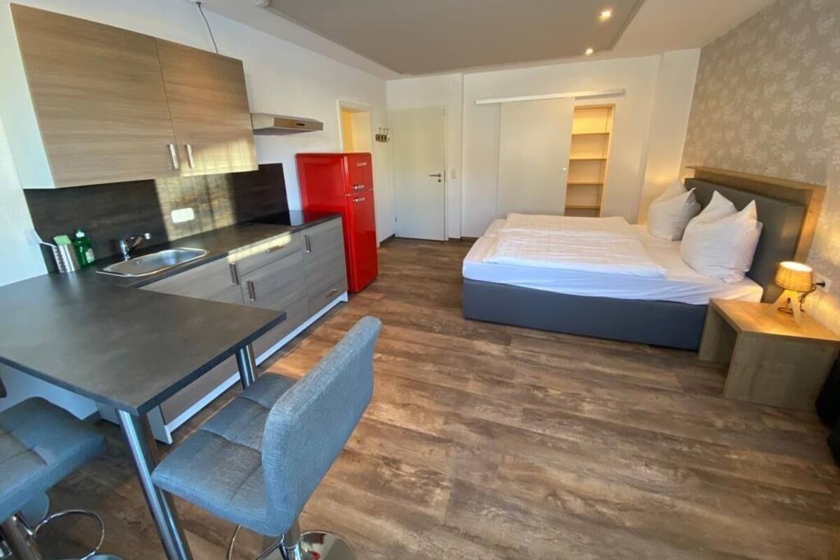 A modern hotel room with a kitchen area, a dining table with chairs, a large bed, and a bedside table with a lamp.