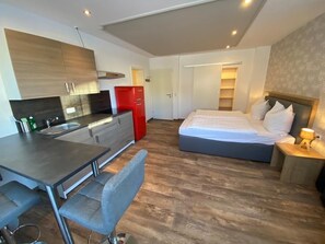 A modern hotel room with a kitchen area, a dining table with chairs, a large bed, and a bedside table with a lamp. - Bergquell Tirol (Jungholz)