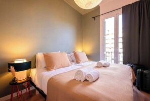A hotel room with a bed, pillows, a nightstand with a lamp, a suitcase, and a window with curtains.