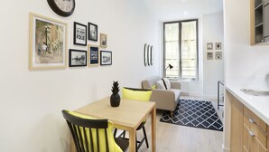 A modern living room with a dining area, featuring a wooden table, chairs, a sofa, and a large window. - Blanche 1 - Studio Apartment, Sleeps 2 (Paris)