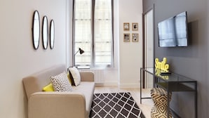 A modern living room with a sofa, a coffee table, a TV mounted on the wall, and a window with blinds. - Blanche 1 - Studio Apartment, Sleeps 2 (Paris)