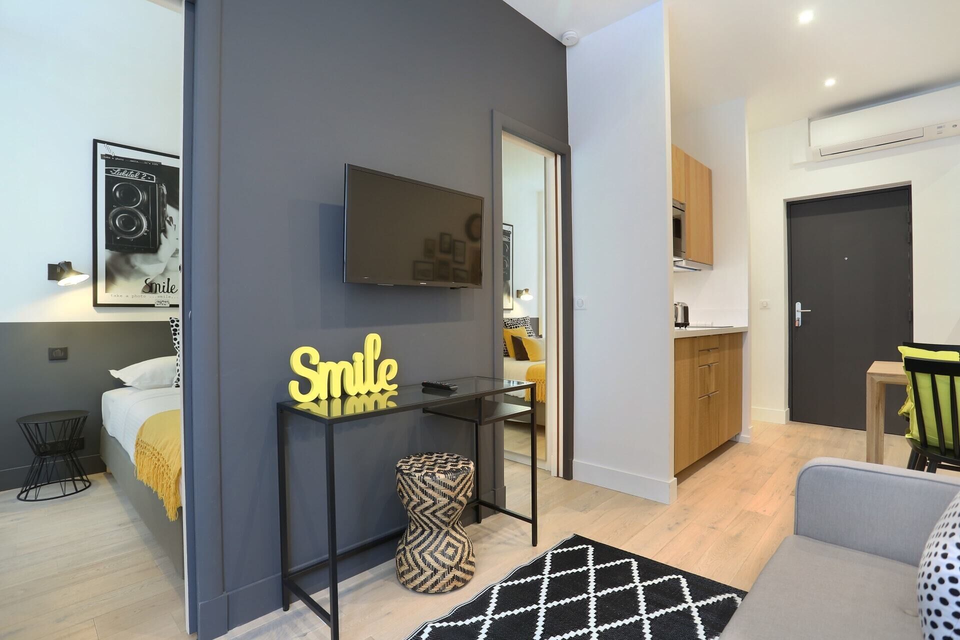 A modern apartment with a grey accent wall, a flat-screen TV mounted above a glass table, a bed with a yellow throw, a kitchen area, and a sofa.