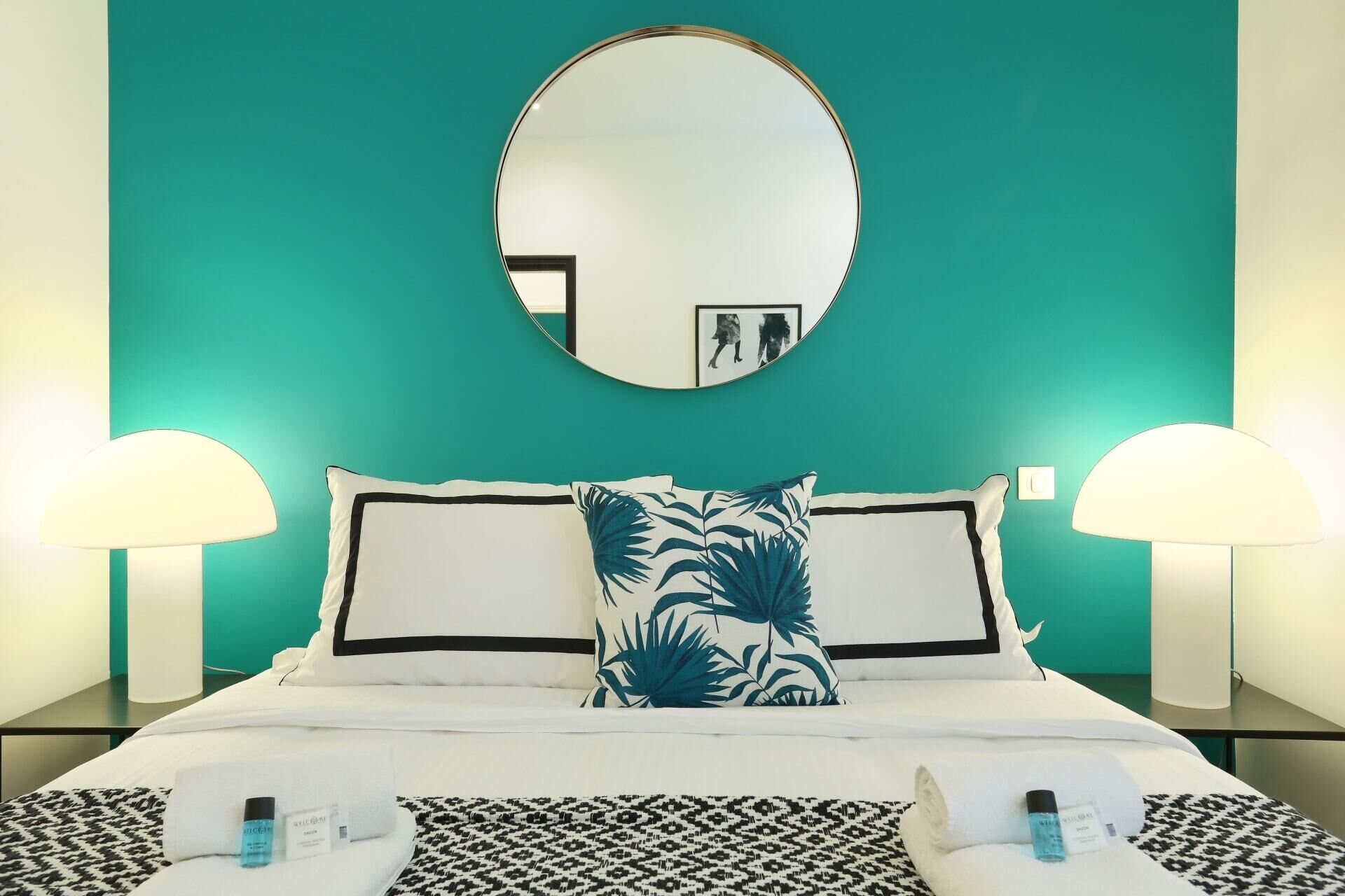 A hotel room with a teal wall, a round mirror, two bedside lamps, a bed with white bedding, a patterned pillow, and a black and white patterned bedspread.