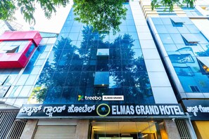The exterior of the Elmas Grand Hotel with a modern glass facade reflecting trees and a red structure to the left. - Treebo Elmas Grand (Bengaluru)