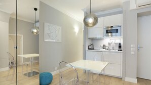 Een moderne keuken met een magnetron, broodrooster en waterkoker. Er is een glazen scheiding tussen de keuken en de eethoek, met een witte tafel en stoelen.