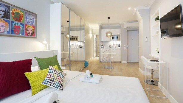 Een moderne hotelkamer met een groot bed, een televisie, een bureau en een keukenhoek zichtbaar door een open deur.