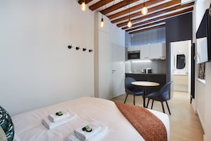 Ein modernes Hotelzimmer mit einem Bett, zwei Sesseln, einem kleinen Tisch und einer Küchenzeile im Hintergrund.