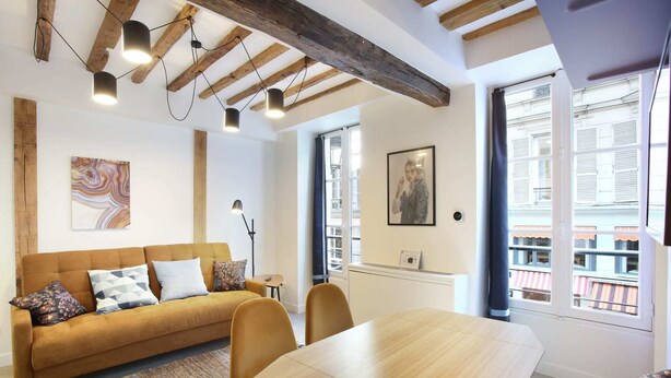 A modern living room with a sofa, a dining table, and a large window with a view of buildings. - Cadet 104 - One Bedroom Apartment, Sleeps 4 (Paris)