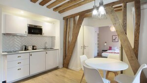 A modern kitchen with white cabinets, a microwave, and a herringbone-patterned backsplash. There is a dining area with a white table and chairs, and a wooden ceiling with exposed beams.
