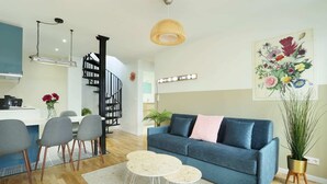 Uma sala de estar moderna com um sofá azul, cadeiras cinzentas, uma mesinha de centro, uma escadaria e uma cozinha ao fundo.