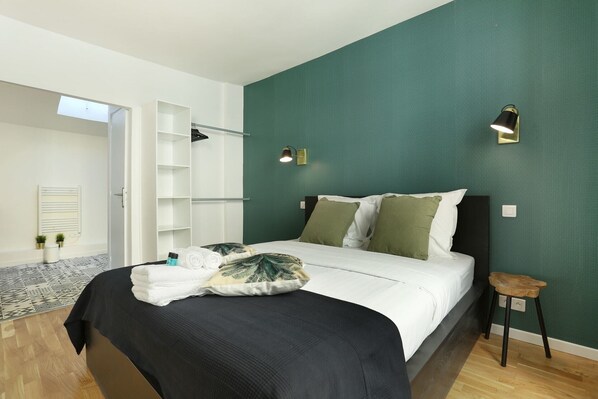 A bedroom with a bed, a nightstand, a wall-mounted lamp, a radiator, and a door leading to another room. - Caire 1 - Two Bedroom Apartment, Sleeps 6 (Paris)